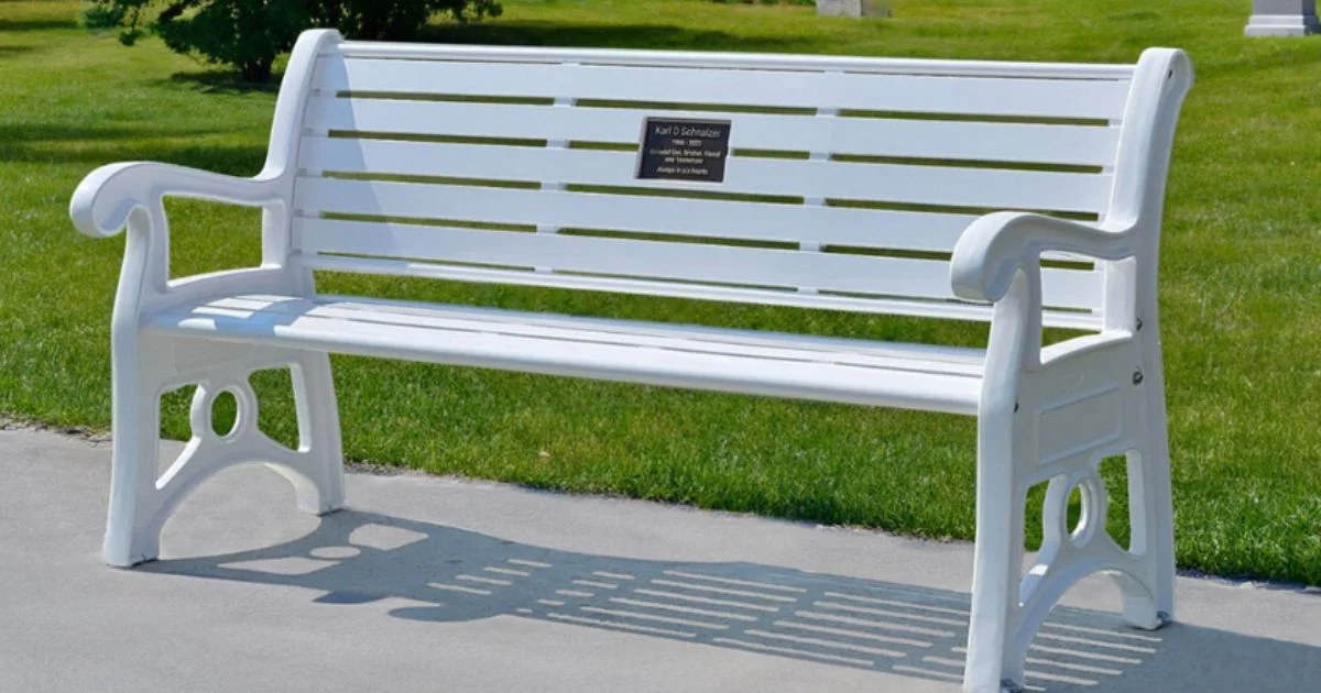 White memorial bench with a dedication plaque, placed on a cemetery path.